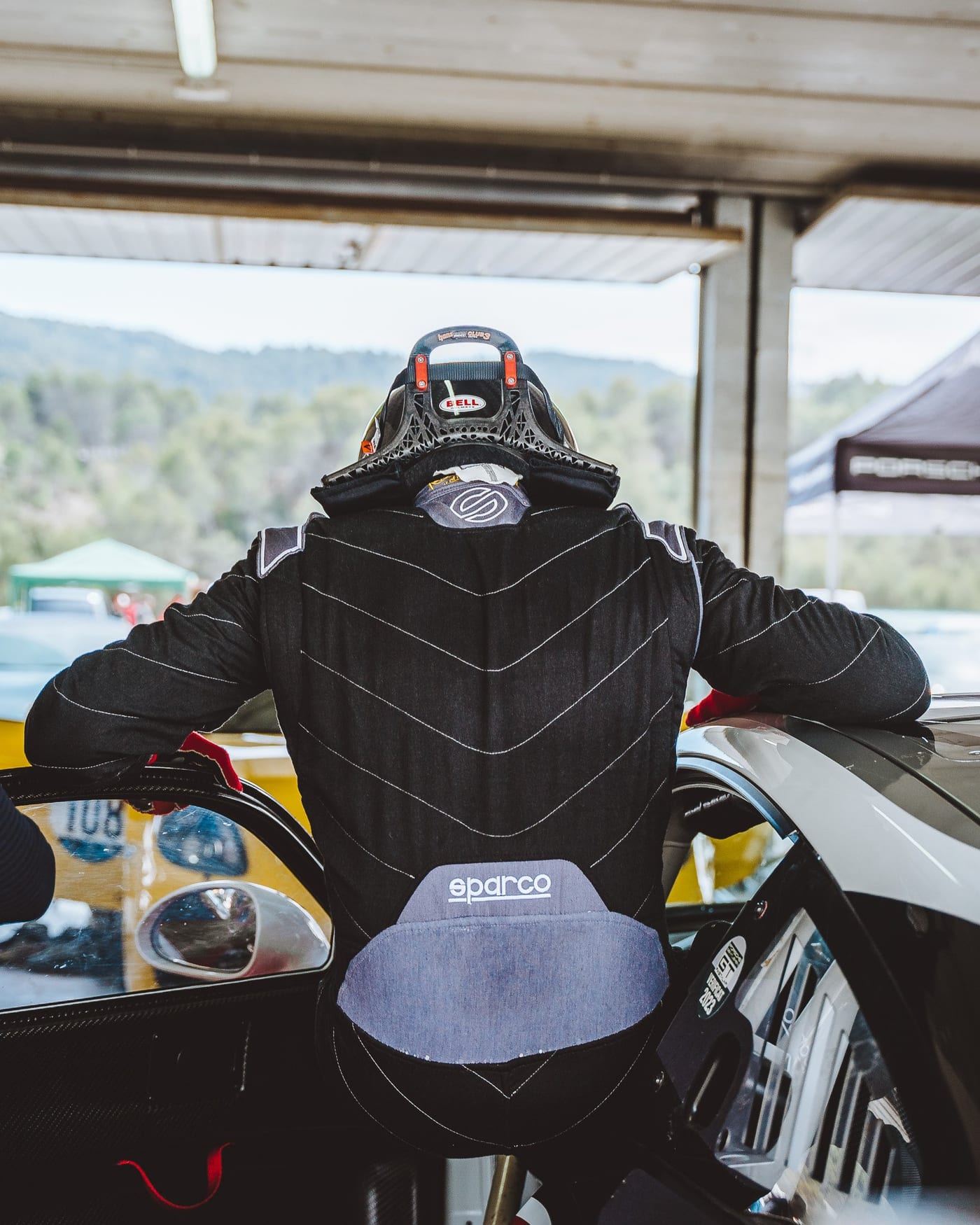 Un piloto con el mono y el casco apoyado en el coche, de espaldas
