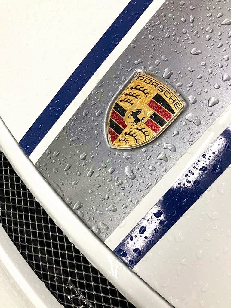 Detall de l'escut Porsche i les bandes blaves d'un capó mullat per la pluja