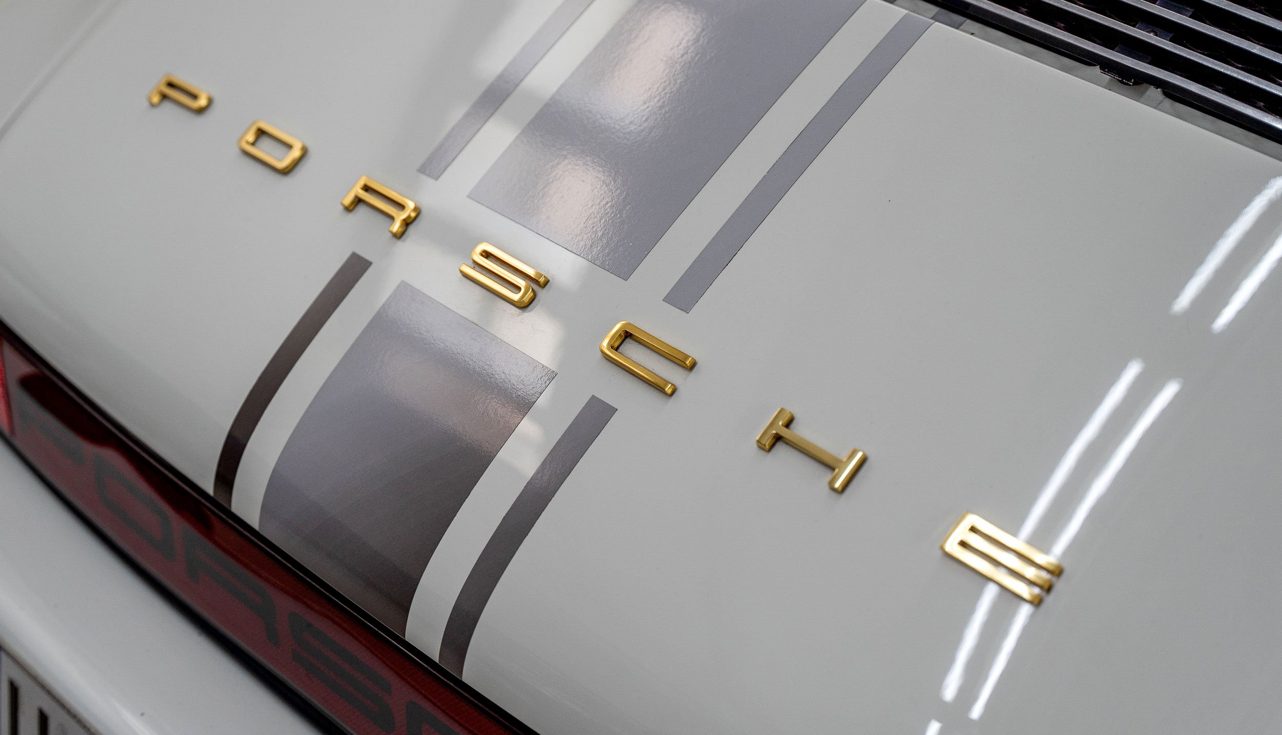 Detall de les lletres daurades de Porsche sobre la tapa del motor blanca amb franges grises